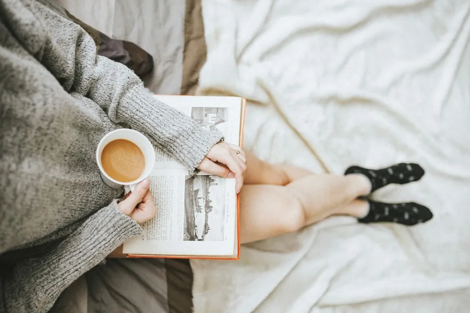 A woman in a grey sweater reading a book with a cup of coffee on her bed