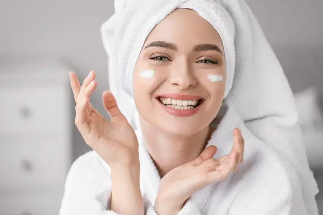 A woman in a bathrobe and hair towel smiling, with cream on her cheeks