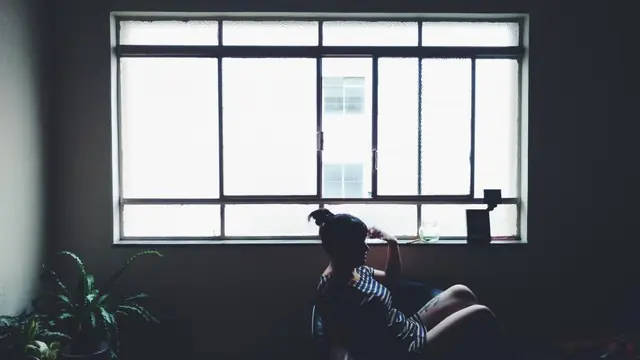 A woman sitting alone in a dim room, looking out of a large window