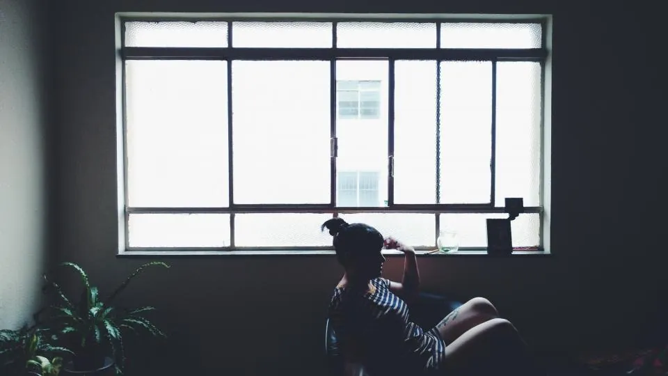 A woman sitting alone in a dim room, looking out of a large window