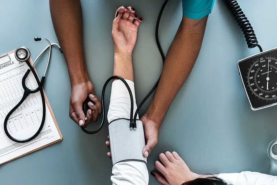 Seen from above, a health worker fitting a blood pressure cuff on a patient's arm, next to a stethoscope, a clipboard and a dial gauge