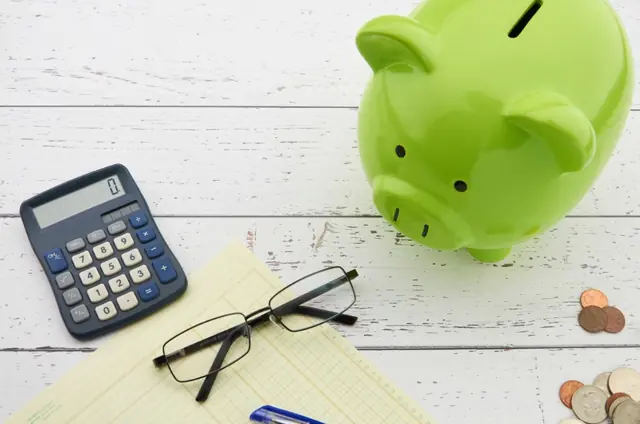 A green piggy bank next to a calculator, glasses, a ledger and coins