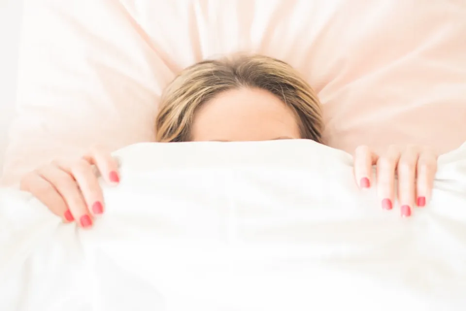 A woman pulling a white duvet up over her face in bed