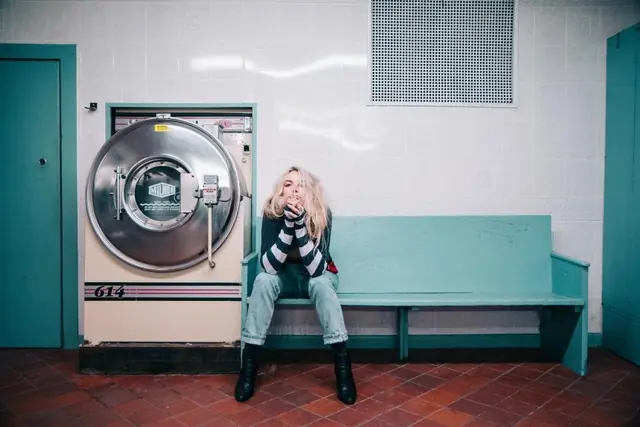 A woman sitting on a bench next to an industrial washing machine in a laundromat