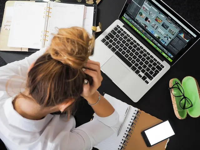 A woman holding her head in her hands at a desk with a laptop and a notebook